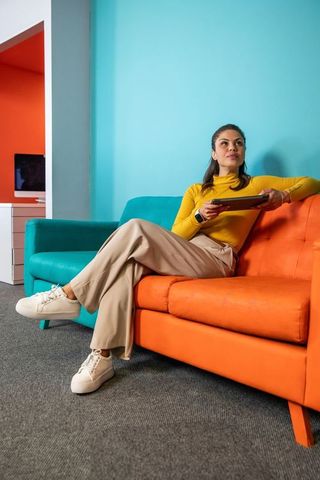 Asian woman sitting on orange sofa holding tablet in vibrant teal coworking lounge