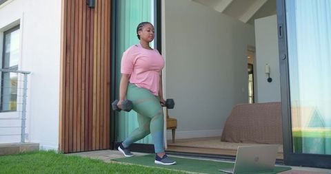African american woman performing dumbbell lunges on home patio with laptop workout