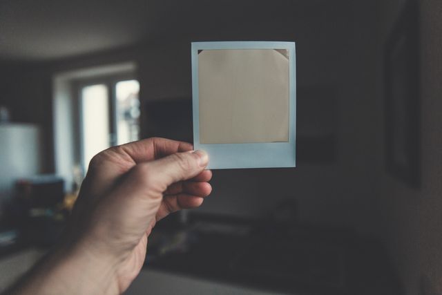 Hand holding blank Polaroid frame in well-lit room. Perfect for concepts of nostalgia, photography, film photography, or inserting your own images into the Polaroid frame.