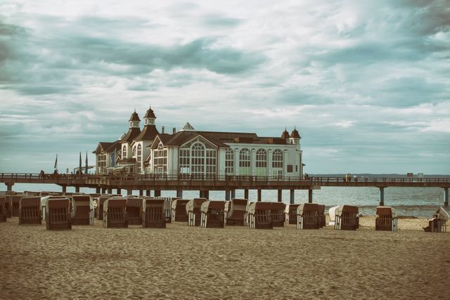 The photo shows a historic pier building extending over the ocean under a cloudy sky. Numerous beach chairs are arranged on the sandy shore, emphasizing a coastal and vintage atmosphere. Good for travel brochures, beach resort advertisements, and inspirational coastal living content.