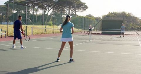 Mixed doubles playing on sunlit outdoor hard court in park with practice wall and fencing