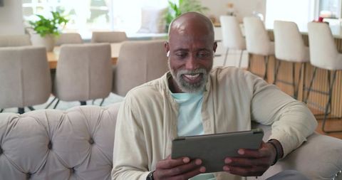 Senior african american man relaxing on tufted sofa using tablet with earbuds in home