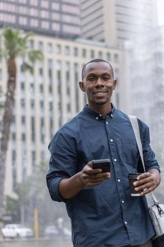 Smiling african man holding smartphone and coffee by city fountain, urban commuter portrait