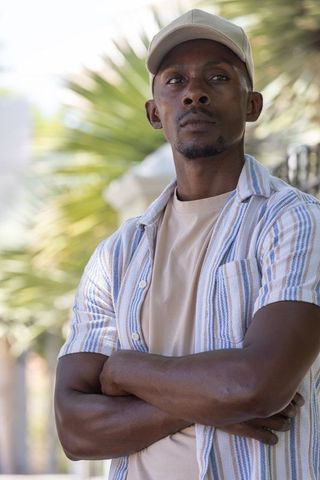 Contemplative african american man standing arms crossed in sunlit garden wearing cap