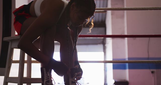 Side view of a female boxer tying her shoes in a gym boxing ring. Captures the moment of preparation and focus. Suitable for use in sports and fitness promotional materials, articles on boxing training, and motivational content.