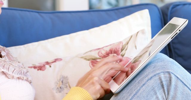 Close-up of a woman using a tablet while sitting on a couch with a floral pillow in background. Suitable for illustrating technology use, casual internet browsing, or modern lifestyles in home settings. Ideal for website banners, tech blogs, or social media content.