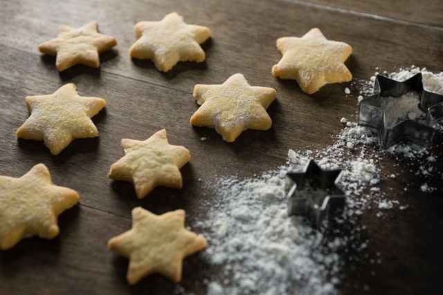 Star-shaped gingerbread cookies sprinkled with powdered sugar on a wooden table. Metal cookie cutters and flour are visible, suggesting a baking scene. Ideal for holiday-themed content, baking blogs, recipe websites, and festive greeting cards.