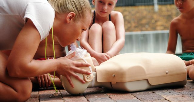 A young Caucasian girl is practicing CPR on a training mannequin, with copy space. She is being observed by two children, highlighting the importance of learning lifesaving techniques from an early age.