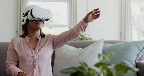 Woman using VR headset on couch reaching toward virtual object in bright modern living room