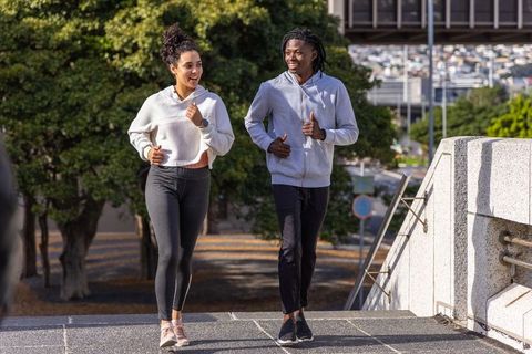 Diverse couple jogging up urban steps wearing hoodies and fitness trackers, smiling