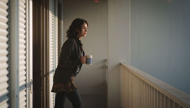 Young woman leaning on balcony railing holding ceramic mug in golden hour light