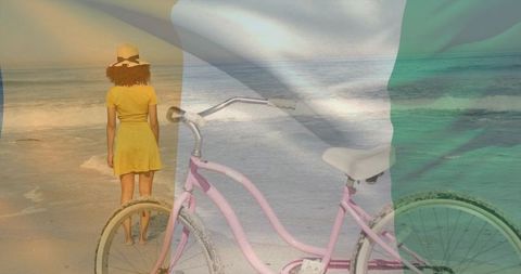 Woman standing facing ocean shoreline with pink bicycle, straw hat and flag overlay during golden ho