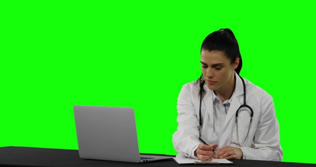 Doctor sitting at desk, recording patient notes on notepad. Laptop open in front. Useful for illustrating healthcare professionals, telemedicine, telehealth services, and remote consultations in medical settings. Green screen background allows for easy integration into various settings or branding scenes.