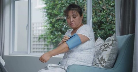 Korean man monitoring blood pressure at home with upper-arm cuff in sunlit living room