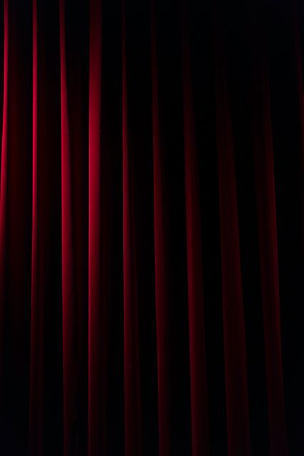Red velvet stage curtains hanging in dim lighting. This can be used for themes related to theater, performances, drama, and elegance. Suitable for promotional material for theatrical productions, artistic events, event posters, and dramatic themed designs.