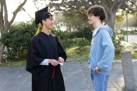 Graduate holding diploma while friend listening on campus path during golden hour