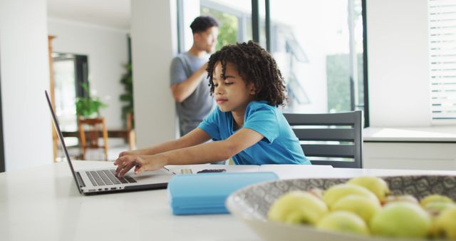 This image features a young boy concentrated on his homework, using a laptop at the kitchen table, while a parent is seen using a smartphone in the background. This scene can be used to depict modern family life, homeschooling, educational technology, and multitasking at home. Suitable for articles or advertisements on parenting, education, and technology use in families.