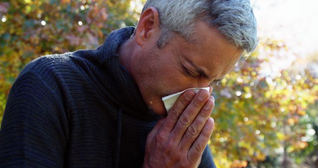 Middle-aged man sneezing into tissue while standing outdoors during autumn. The man appears to suffer from allergies or a cold. The changing colors of autumn leaves are visible in the background, emphasizing the outdoor setting. This image is suitable for use in articles or advertisements related to allergies, seasonal illnesses, health care, and outdoor activities in the fall.