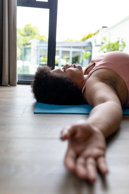 African American woman lying on yoga mat with arms outstretched, practicing relaxation and mindfulness at home. Ideal for use in articles or advertisements related to fitness, wellness, home workouts, self-care routines, and promoting a healthy lifestyle.