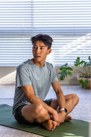 Asian man practicing mindful stretching on green yoga mat at home wearing smartwatch