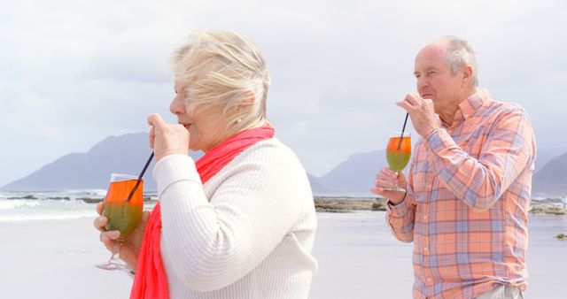 Senior couple enjoying refreshing cocktails on the beach with scenic mountains and ocean in the background. Ideal for use in travel, leisure, retirement, and healthy living promotions.