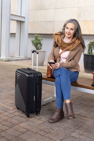 Mature woman waiting at transit plaza with suitcase using smartphone, urban travel