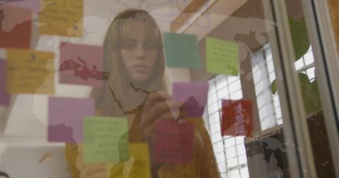 Woman placing sticky notes on glass while reviewing global business plan and brainstorming