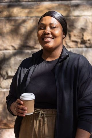Sunlit african woman smiling and holding coffee cup in urban courtyard with stone wall