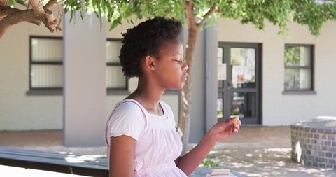 Teenage girl sitting on bench enjoying snack while studying under leafy campus tree