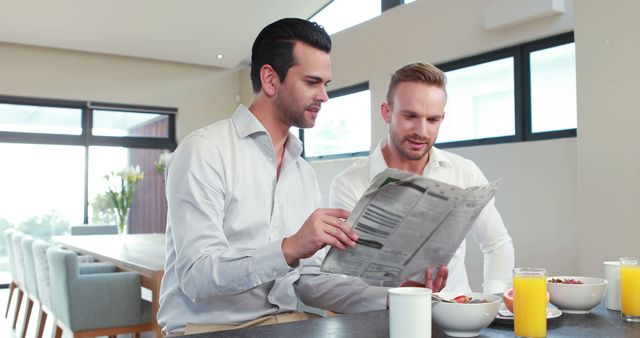 Two Caucasian men are engaged in reading a newspaper together during breakfast in a modern kitchen, with copy space. Their casual attire and relaxed posture suggest a comfortable, domestic setting.