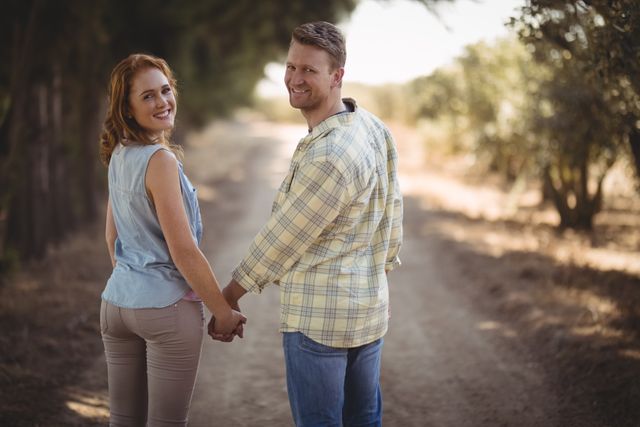 Young couple holding hands while walking on a dirt road at an olive farm. Both are smiling and dressed in casual clothing, enjoying a peaceful moment in nature. Ideal for use in advertisements, relationship blogs, travel brochures, and lifestyle magazines.