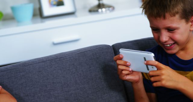 Young boy sits on sofa engrossed in a game on his smartphone. He is smiling, showing enjoyment and focus. Perfect image for articles or ads about children and technology, games, social interactions, or indoor leisure activities.