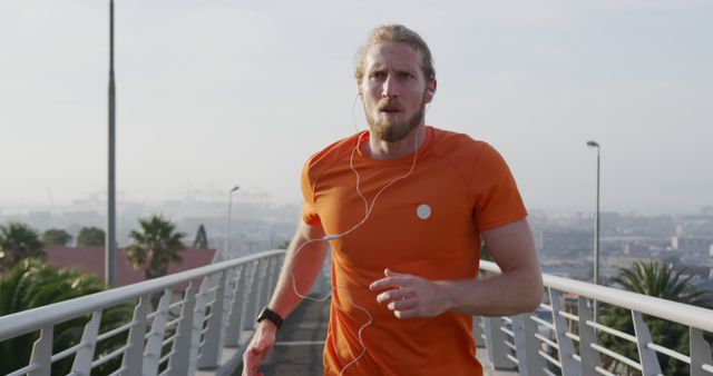 Blonde man jogging on urban bridge wearing orange shirt and earbuds. Cityscape and palm trees in background. Great for fitness promotions, outdoor exercise themes, urban lifestyle content, and healthy living campaigns.