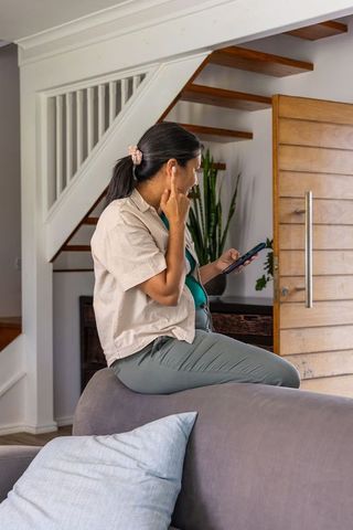 Mature asian woman adjusting earbud while holding smartphone sitting on sofa back by front door
