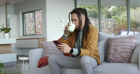 Man sipping coffee and checking smartphone on modern living room sofa