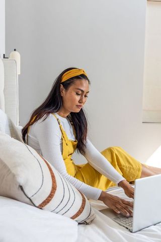Asian woman typing on laptop in cozy bedroom wearing mustard overalls and headband