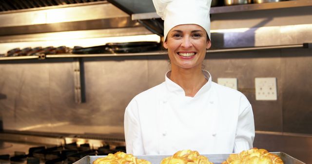 Female chef holding a tray of freshly baked pastries in a commercial kitchen while smiling. Ideal for use in advertisements for cooking classes, culinary schools, bakery promotions, or restaurant and catering services showcasing skilled chefs and freshly baked goods.