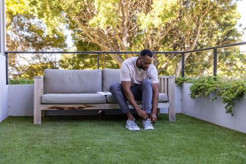 Mature african american man tying sneakers on rooftop terrace sofa with tablet nearby