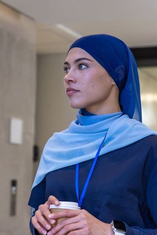Woman standing wearing two-tone blue headscarf holding coffee in lobby