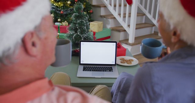 An elderly couple wearing Santa hats enjoys a virtual celebration seated before a decorated Christmas tree. They are holding coffee mugs and have a plate of cookies on the table. This cheerful holiday scene can be used for promotions or articles related to virtual celebrations, family connections during holidays, or advertisements for holiday gifts and online services.