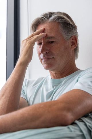 Pensive senior man resting on sofa by window reflecting in natural daylight