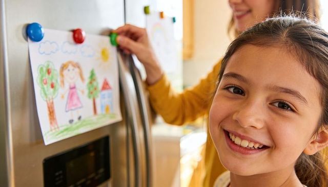 Smiling school-age girl displaying refrigerator artwork while parent secures colorful magnet