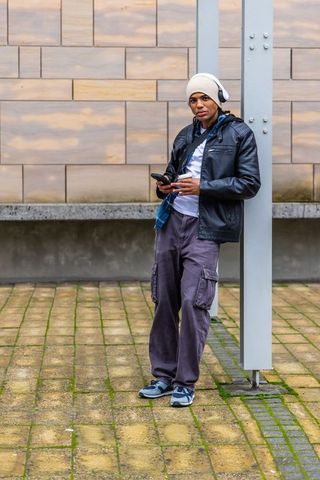 Young man leaning on column listening to music using smartphone in urban courtyard