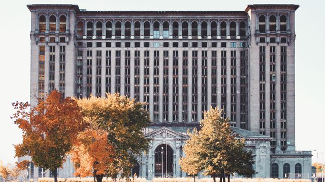 Image features an abandoned historic train station with towering architecture situated against an autumn backdrop. Trees with colorful foliage enhancing the foreground of the weathered, decades-old structure. Perfect for illustrating urban decay, historical architecture, or transformation of cityscapes over time. Use for projects on historic landmarks, the impact of the passage of time, or the appeal of autumn's natural beauty surrounding urban settings.