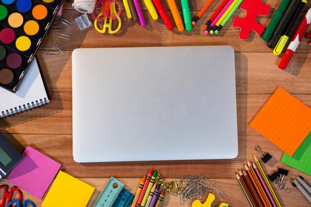 A laptop is closed and surrounded by a variety of colorful stationery items such as pencils, markers, paper clips, sticky notes, and a notepad on a wooden desk. This image is great for illustrating creative workspaces, educational materials, home office setups, and organizational themes.