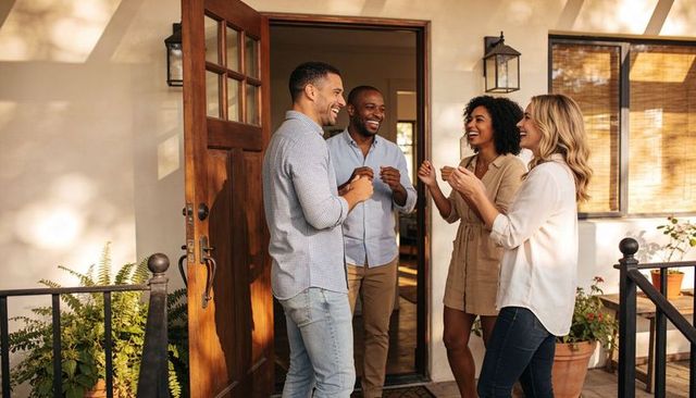 Laughing diverse friends greeting on sunny front porch with wooden door and potted plants