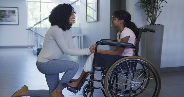 This image shows a caring woman kneeling beside a young girl who is in a wheelchair, with both smiling and interacting warmly indoors. The setting appears modern and spacious, possibly a healthcare facility or an educational institution. Ideal for use in promoting healthcare services, mental health awareness, inclusivity, support groups, and caregiving services.