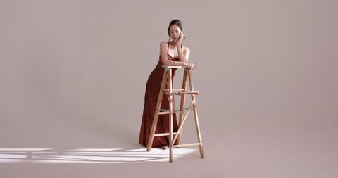 Elegant woman in rust gown leaning on ladder in minimalist studio