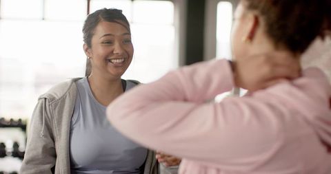 Multiracial women chatting in bright gym near dumbbells, smiling post-workout camaraderie