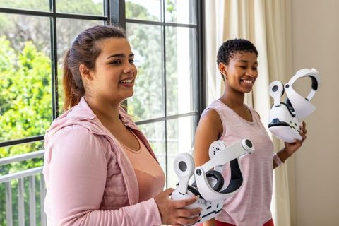 Multicultural women holding vr headsets by sunlit window preparing virtual reality session